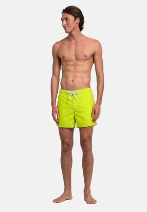 Uomo senza maglietta in piedi a piedi nudi che indossa pantaloncini da bagno giallo brillante con coulisse bianca, guarda a sinistra su sfondo uniforme.