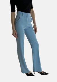 Lichtblauwe, op maat gemaakte broek met rechte pijpen, hoge taille, steekzakken aan de voorkant en knoopsluiting, gecombineerd met zwarte puntige hakken.