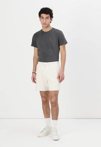 T-shirt in cotone grigio con taschino sul petto, abbinato a pantaloni corti elasticizzati bianchi. Il modello indossa calze bianche e sneakers bianche. Sfondo semplice.