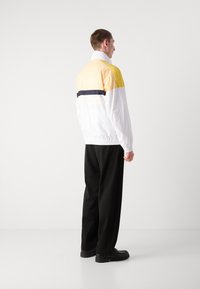 Jachetă din material impermeabil cu blocuri de culoare alb și galben, având o dunga orizontală bleumarin, cu manșete elastice și un croi lejer, purtată cu pantaloni negri.