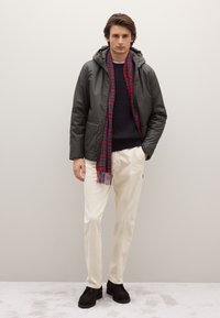 Giacca impermeabile verde scuro con cappuccio, maglione lavorato a maglia blu navy, pantaloni color crema e sciarpa a quadri rossa, abbinati a scarpe marrone scuro.