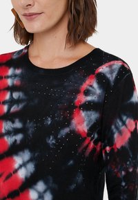 Haut en maille tie-dye noir, rouge et bleu avec encolure ronde, orné de petits clous métalliques, texture lisse, et manches longues.