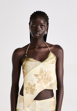 Gelbes und cremefarbenes Neckholder-Top mit asymmetrischem Schnitt, verziert mit floralen Mustern und weichem Material. Geflochtenes Haar und Ohrringe runden den Look ab.
