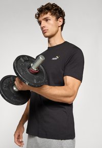 Zwart katoenen t-shirt met korte mouwen en een klein gouden Puma-logo. De persoon houdt een zwart gewicht vast met een zilveren handgreep.