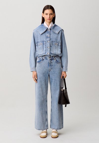 Giacca di jeans a lavaggio chiaro con colletto ampio e tasche sul petto, abbinata a jeans a gamba larga. Il modello tiene in mano una piccola borsa nera, indossando scarpe bianche.