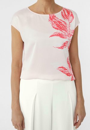 Femme portant un haut à manches courtes rose clair avec un motif floral rouge sur le côté droit, associé à un pantalon blanc taille haute.