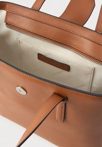 Brown leather tote bag with a smooth texture, featuring an interior zipper pocket and branded tag. Lined with beige material.