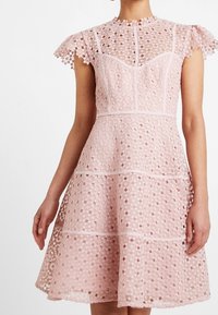 Vaaleanpunainen pitsimekko, jossa on hihattomat hihat, muotoiltu yläosa ja levenevä hame. Sisältää geometrisia leikkuja, kontrastoivia vuoria ja vaakasuuntaisia pitsikuvioita.