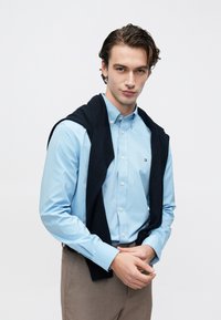 Chemise bleu clair boutonnée avec manches retroussées et petit logo, associée à un pull bleu marine noué autour du cou, et un pantalon beige.