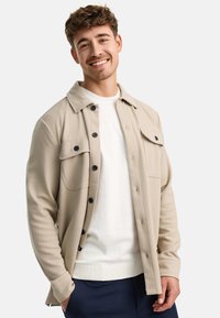 Chaqueta de camisa beige con cierre de botones y bolsillos en el pecho, usada sobre un suéter blanco. Presenta un tejido texturizado y botones oscuros.