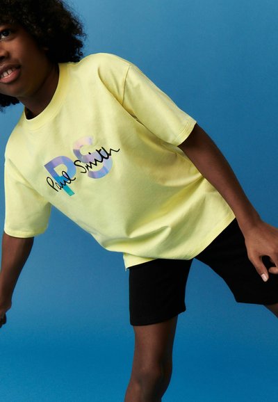 Camiseta de algodón amarilla con "PS" en un estilo holográfico y "Paul Smith" en cursiva negra, combinada con pantalones cortos negros. Fondo azul.
