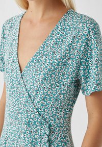 Femme portant une robe enveloppante teal avec un petit motif floral blanc et noir, à manches courtes, coupée aux épaules et à la taille.