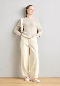 Donna con lunghe trecce in coda di cavallo, indossa un maglione di lana beige, pantaloni a gamba larga crema, scarpe nude e porta una borsa a spalla beige, in piedi all'interno.