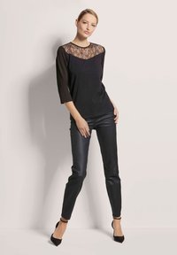 Zwarte top met kanten inzetstuk op de schouders en driekwart mouwen, gecombineerd met zwarte strakke broek en hoge hakken, tegen een neutrale achtergrond.