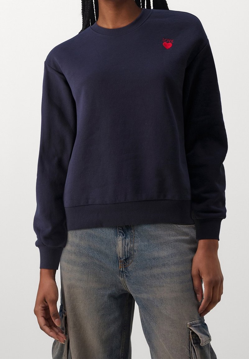 Femme portant un sweat-shirt bleu marine avec un petit cœur rouge et le texte "LOVE" sur la poitrine, associé à un jean cargo bleu clair.