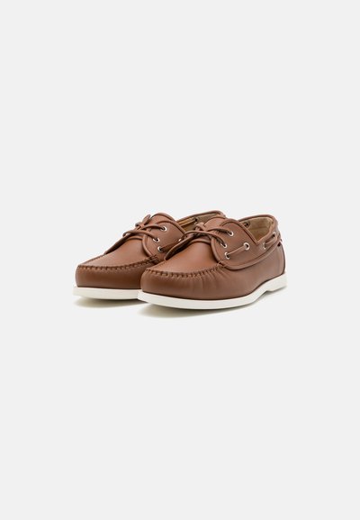 Pantofi de barcă din piele maro cu talpă din cauciuc alb, vârf rotunjit, cusături contrastante și gromete metalice pentru șireturi. Design casual.