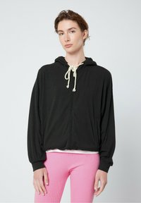 Melns zip-up džemperis, viegla auduma, ar kapuci un priekšējiem kabatām. Saskaņots ar rozā ribotām biksēm.