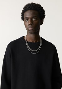 Pulover negru cu decolteu rotund, având un tricot texturat. Două lanțuri argintii stratificate ca accesorii, plasate pe un fundal deschis.