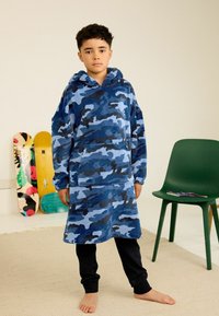 Ein Kind trägt ein himmelblaues Camouflage-Hoodie-Kleid über schwarzen Hosen und steht barfuß in einem Raum, in dem zwei Skateboards und ein grüner Stuhl in der Nähe sind.