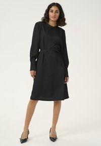 Robe midi noire avec des manches longues, un col rond et une ceinture en tissu à la taille. Texture lisse, silhouette simple et sans motifs visibles.
