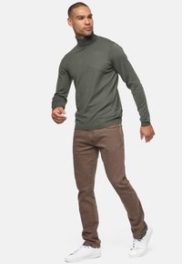 Jeans de mezclilla marrones combinados con un suéter de cuello alto verde oscuro. El modelo lleva zapatillas blancas. Tejido suave con silueta ajustada y cuello de tortuga acanalado.