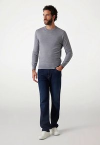 Šedý sveter s dlhými rukávmi a okrúhlym výstrihom, v kombinácii s tmavomodrými džínsami a bielymi teniskami. Model stojí nenútene s rukami v vreckách na neutrálnom pozadí.