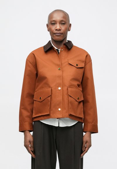 Brown cotton jacket with a collar, two front patch pockets, gold hardware accents, and a structured fit. Worn over a white shirt.