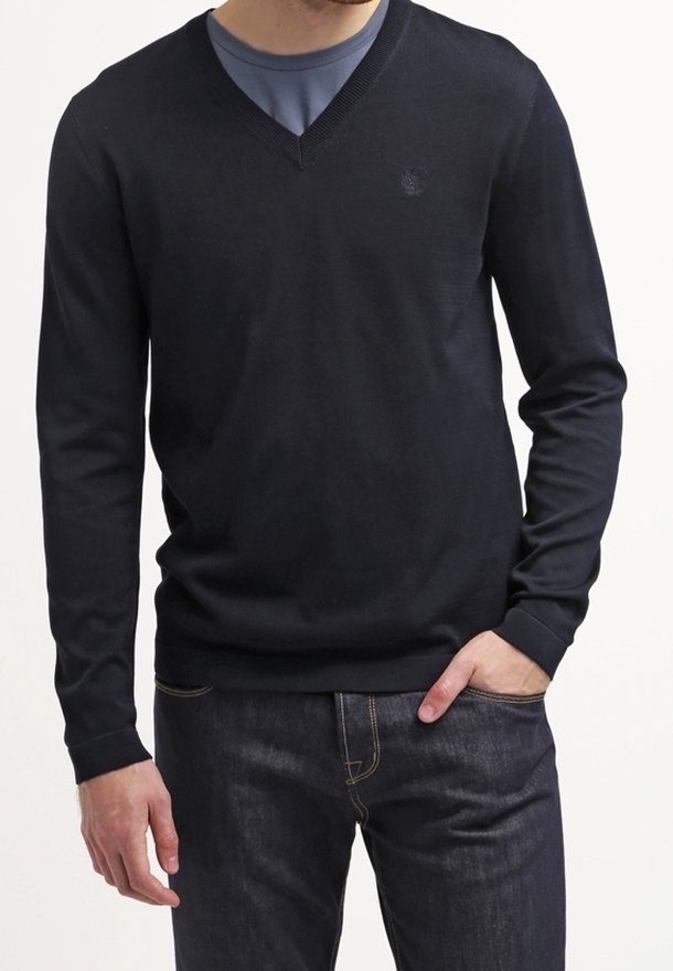 Osoba oblečená v tmavém svetr s výstřihem do V přes modrou košili, s rukama v kapsách, stojí v klidu na světlejším pozadí.