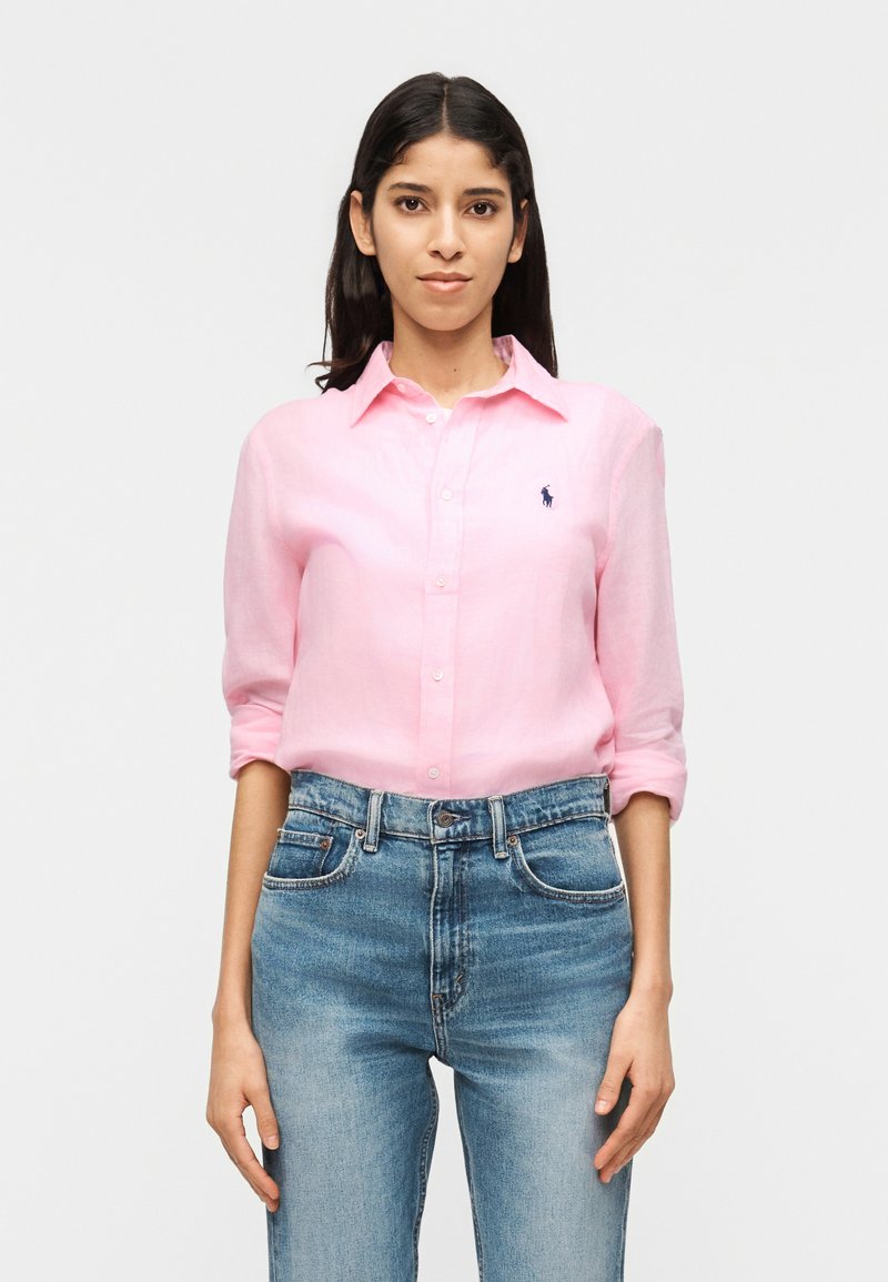 Femme aux longs cheveux foncés portant une chemise rose clair à boutons avec les manches retroussées et un jean bleu taille haute, debout devant un fond blanc.