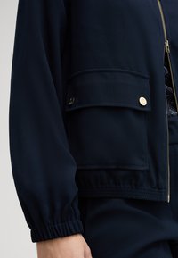Dunkelblaue Jacke mit Reißverschluss vorne, elastischen Bündchen und einer aufgesetzten Tasche mit goldenen Hardware-Details. Hergestellt aus einem strukturierten Stoff.