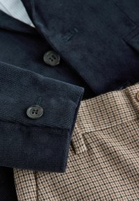 Námornícka kordová bunda s čiernymi gombíkmi skombinovaná s nohavicami s houndstooth vzorom v hnedej farbe, ktorá vystihuje textúrovanú látku a prispôsobený dizajn.
