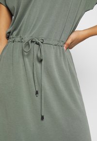 Primer plano de una persona ajustando el cordón de la cintura de un vestido casual verde claro contra un fondo claro y liso.