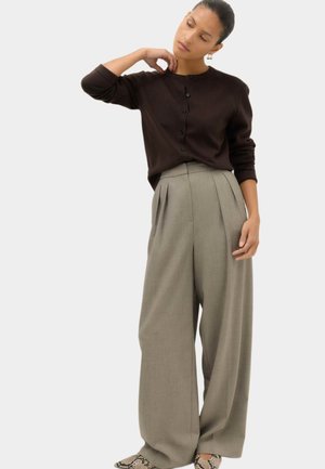 Femme aux cheveux attachés, portant un cardigan noir boutonné, un pantalon gris large et des chaussures à motif peau de serpent, debout avec une main sur le cou.
