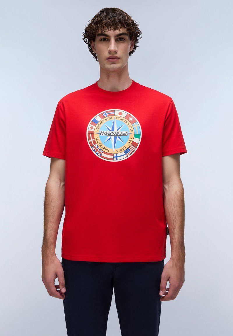 Rød bomulds t-shirt med et cirkulært grafisk motiv, der har en blå stjerne og flere nationale flag. Kortærmet og med rund hals.