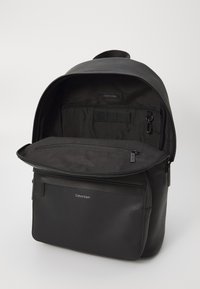Zaino nero realizzato in materiale testurizzato, con parte superiore arrotondata, una tasca frontale con zip e un interno dotato di scomparti organizzativi.