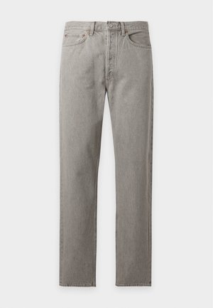 Jeune homme debout portant une chemise rouge foncé à manches longues, un jean gris clair et des chaussures noires sur un fond clair uni.