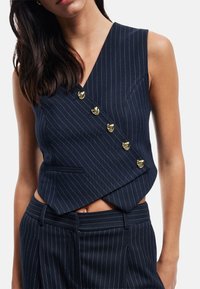 Marineblaues, gestreiftes ärmelloses Top mit V-Ausschnitt, das goldene Knöpfe und eine Seitentasche bietet. Der Stoff hat eine glatte Textur und ein tailliertes Design.