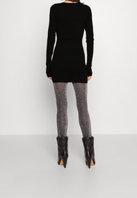 Svart långärmad klänning, figurnära vid midjan, ihop med glittriga genomskinliga leggings och svarta klackstövlar, visad mot en enkel bakgrund.