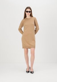 Abito maglione beige, maniche lunghe, scollo rotondo, spacchi laterali, abbinato a tacchi alti neri e occhiali da sole, design minimalista.
