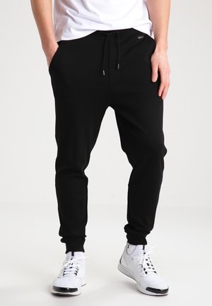 Mand iført sorte joggerbukser med snøre og hvide sneakers, stående med hænderne i lommerne på hvid baggrund.