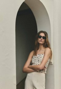 Femme portant des lunettes de soleil noires, un haut sans manches à pois et un pantalon clair, appuyée contre une porte blanche en arc avec les bras croisés.