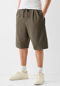 Olivově zelené kraťasy z měkké tkaniny, s elastickým pasem na šňůrku a bočními kapsami, v kombinaci s bílými teniskami.