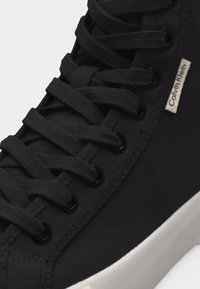 Μαύρο αθλητικό παπούτσι Calvin Klein με μαύρα κορδόνια, μαύρες οπές για κορδόνια και λευκή σόλα, με κοντινή λήψη στην ετικέτα λογότυπου στο πλάι και την περιοχή των κορδονιών.