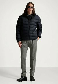 Nuori mies, joka pukeutunut mustaan untuvatakkiin, tummansävyiseen villapaitaan, harmaisiin housuihin, mustiin Chelsea-saappaisiin ja pyöreisiin mustiin aurinkolaseihin, seisoo yksinkertaista taustaa vasten.