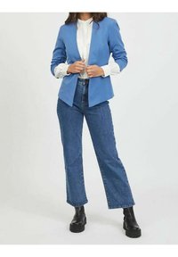 Modrý oblekový blazer s klopami, spárovaný s bílou košilí a vysokými splývavými džíny. Celý vzhled doplňují masivní černé boty.