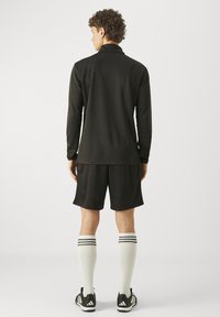 Man met de rug naar de camera, gekleed in een zwart shirt met lange mouwen, zwarte korte broek, witte kniehoge sokken met strepen en zwarte sportschoenen met witte zolen.