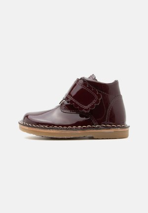 Glänzender dunkelroter Kinderknöchel-Stiefel mit wellenförmigem Klettverschluss-Riemen und beiger Gummisohle auf weißem Hintergrund.