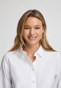 Witte blouse met lange mouwen, voorzien van een klassieke kraag en lichte knopen. Model heeft los, steil haar en glimlacht.