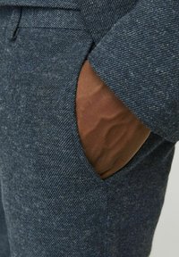 Marineblaue, strukturierte Stoffhosen mit einer Seitentasche, in der eine braune Hand zu sehen ist. Der Stoff weist ein dezentes Fischgrätmuster auf.