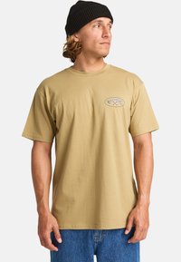Beige katoenen T-shirt met korte mouwen en een ronde hals. Heeft een logopatch op de linker borstpartij. Draag het met blauwe denim short.